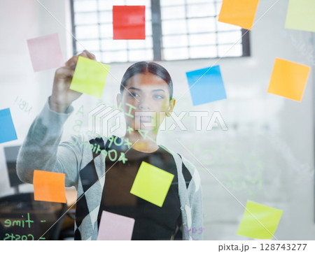 Woman arranging colorful sticky notes on glass board in open-plan office, with monitors and chairs 128743277
