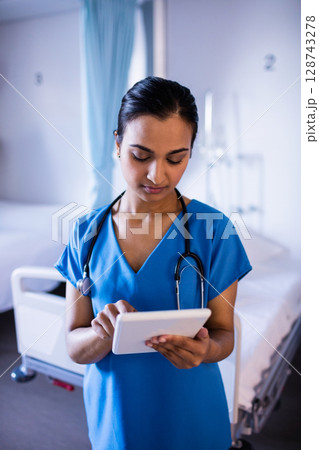 Young Indian nurse standing in hospital room, checking tablet with stethoscope and IV stand 128743278