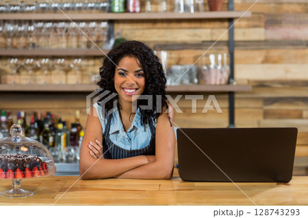 Woman leaning on wooden café counter, showcasing open laptop and glass cake dome with strawberries 128743293