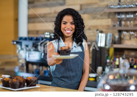 Young adult woman barista serving chocolate muffin on counter in cafe, featuring espresso machine Young adult woman barista serving chocolate muffin on counter in cafe, featuring espresso machine 128743295