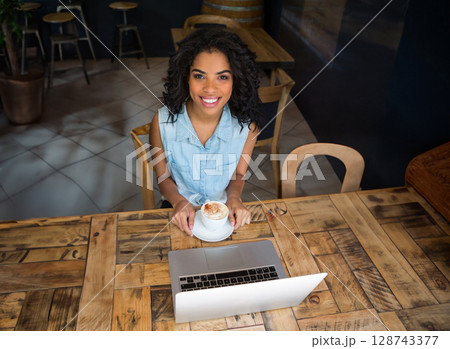 Woman sitting alone at rustic wooden table in café, holding frothy cappuccino cup and using laptop Woman sitting alone at rustic wooden table in café, holding frothy cappuccino cup and using laptop 128743377