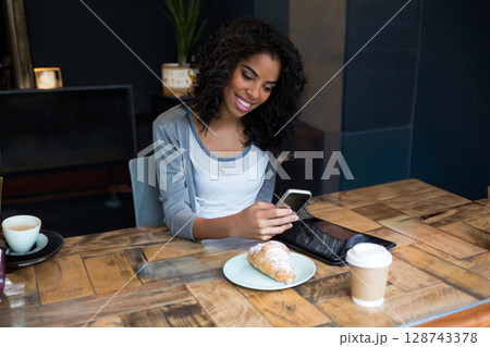 Woman sitting at wooden table in cozy café, using smartphone and tablet with latte and croissant 128743378