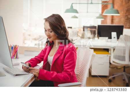 Asian woman using smartphone at white desk in open-plan office, with monitor and colored pencils 128743380