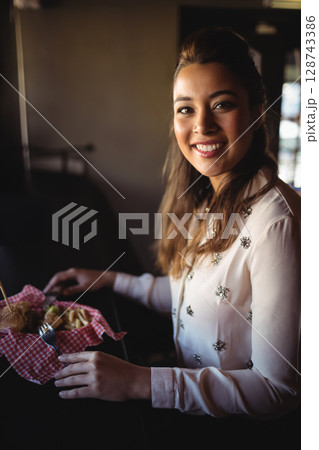 Young woman eating burger and fries in checkered basket at table in cozy café, copy space 128743386