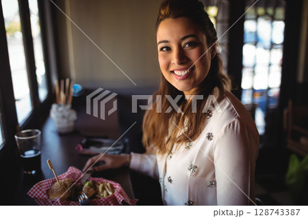 Young woman sitting at counter by window in café, eating fries from checkered basket, sipping drink 128743387