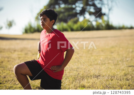 Boy performing lunge stretch on dry grassy field outdoors, with yellow wrist wrap and red T?shirt 128743395