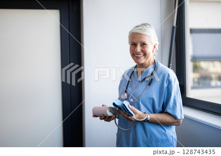Senior female nurse holding blood pressure monitor and cuff at clinic window, copy space 128743435