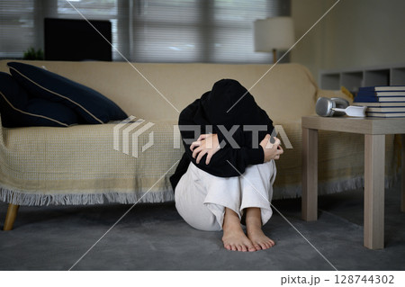 Sad teenage girl sitting alone by the couch with feelings of anxiety, depression, or isolation 128744302