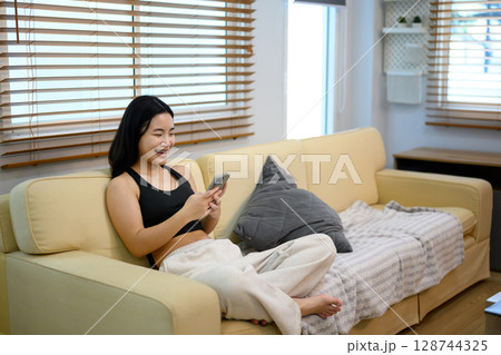 Happy teenage girl relaxing on a sofa, laughing and browsing her smartphone 128744325