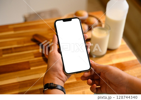 Hands holding mobile phone with empty display over wooden kitchen counter and breakfast 128744340