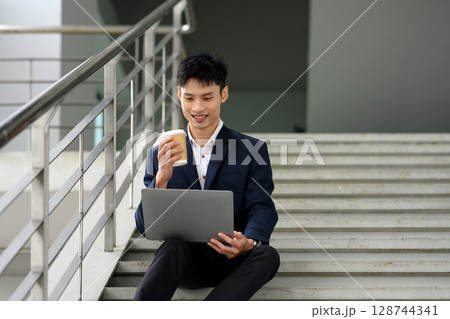 Businessman sitting on stairs sipping takeaway coffee and using laptop 128744341