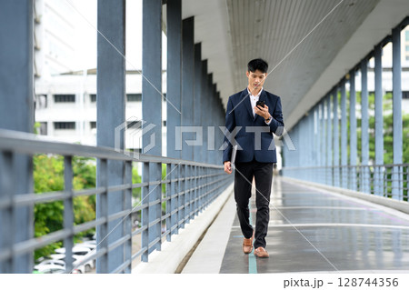Businessman walking confidently on pedestrian bridge while checking smartphone Businessman walking confidently on pedestrian bridge while checking smartphone 128744356