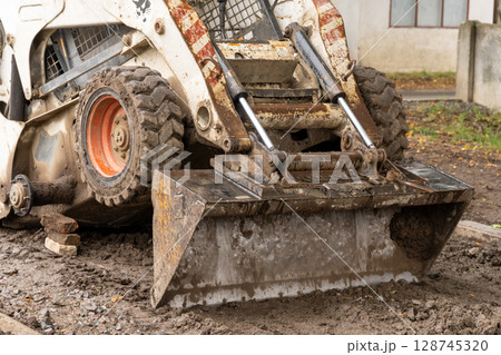 A skid steer loader with a dirt-filled bucket actively works on a muddy construction site, preparing for new building projects A skid steer loader with a dirt-filled bucket actively works on a muddy construction site, preparing for new building projects 128745320