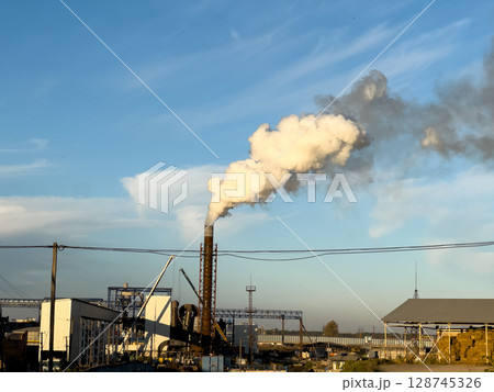 White smoke rises from a tall chimney against a clear blue sky, highlighting industrial activity in an urban environment 128745326