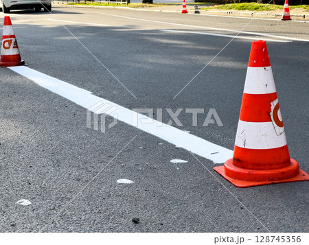 Traffic cones are positioned along a freshly painted lane marking in an urban area during road maintenance work, ensuring safety for drivers 128745356