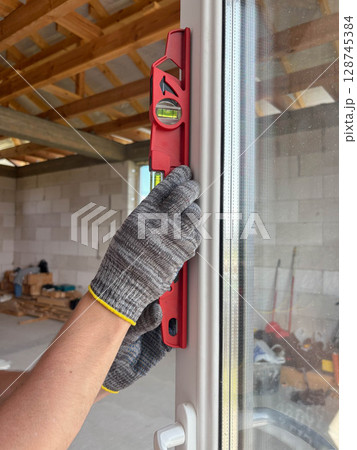 A worker wearing gloves and using a level checks the evenness of a new window at a construction site. 128745384
