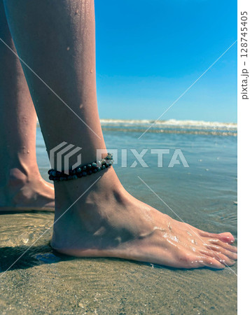 Beautiful female legs with a anklet on the beach. Sea wave washes female legs. 128745405
