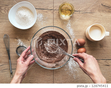 Female hands preparing chocolate sponge cake. Glass bowl with cocoa and flour on wooden table. Female hands preparing chocolate sponge cake. Glass bowl with cocoa and flour on wooden table. 128745411