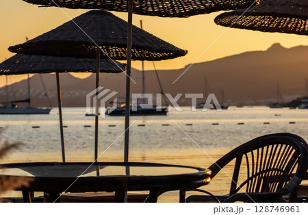 Golden hour light bathes beach umbrellas, outdoor furniture, and feathery reeds, overlooking a tranquil bay with distant mountains, creating a serene sunset scene. High quality photo Golden hour light bathes beach umbrellas, outdoor furniture, and feathery reeds, overlooking a tranquil bay with distant mountains, creating a serene sunset scene. High quality photo 128746961