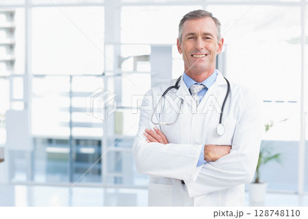 Mature male doctor smiling with stethoscope, crossed arms in clinic beside potted plant, copy space 128748110