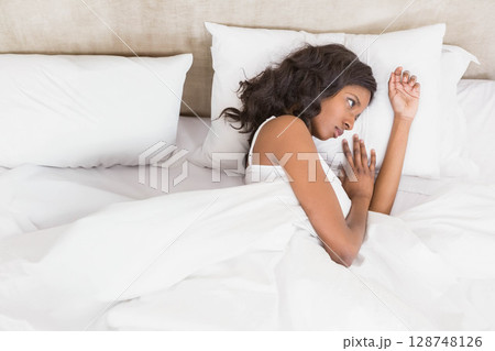 African American woman lying under white bedding on bed in bedroom wearing white tank top African American woman lying under white bedding on bed in bedroom wearing white tank top 128748126