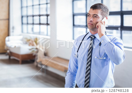 Male doctor wearing shirt and tie talking on smartphone in lobby with stethoscope, copy space 128748128