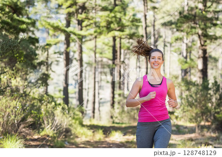 Female runner wearing sportswear jogging on dirt trail through sunlit pine forest, copy space 128748129