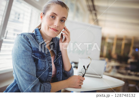 Woman holding smartphone to ear, holding glasses and using laptop at modern office table 128748132
