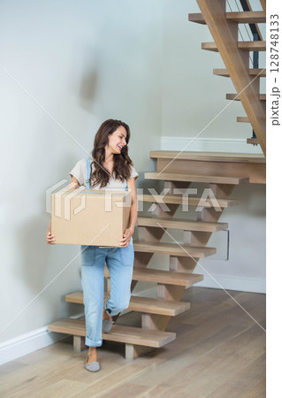 Mid adult woman carrying cardboard moving box while climbing wooden staircase in home stairwell 128748133