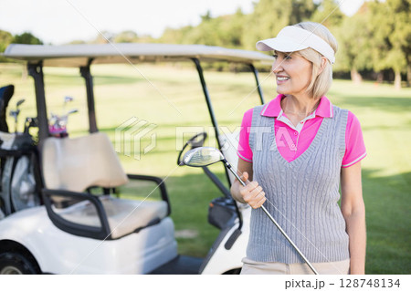 Senior woman wearing pink polo standing on fairway next to golf cart holding driver, copy space 128748134