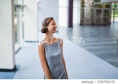 Mid-20s woman standing in modern lobby wearing satin dress by columns, glass doors, and windows Mid-20s woman standing in modern lobby wearing satin dress by columns, glass doors, and windows 128748141