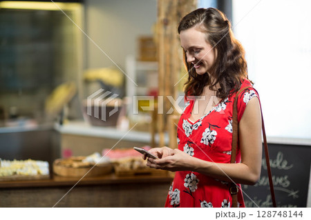 Woman standing at wooden counter in bakery checking smartphone by wicker pastry baskets, copy space 128748144