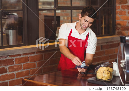 Male barista wearing apron handling payment at cafe counter by payment terminal and credit card Male barista wearing apron handling payment at cafe counter by payment terminal and credit card 128748190