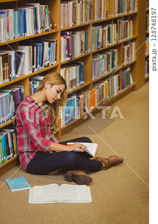 Female student sitting on library aisle turning pages of open book beside closed books copy space 128748197