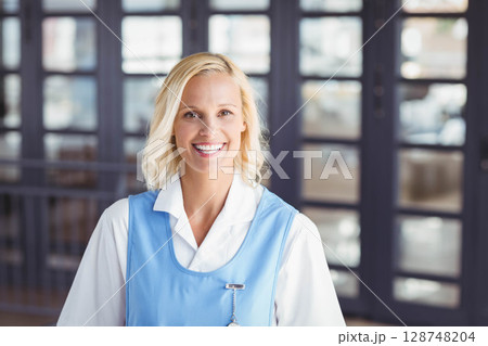Middle-aged female hotel staff member standing in blue smock at lobby doors with name badge Middle-aged female hotel staff member standing in blue smock at lobby doors with name badge 128748204