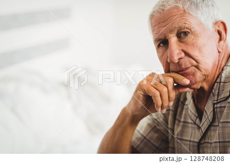 Senior man sitting on bed in bedroom with white headboard, resting chin on hand, copy space 128748208