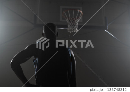 Basketball player standing at free-throw line in gym, looking up at hoop mounted on metal beams Basketball player standing at free-throw line in gym, looking up at hoop mounted on metal beams 128748212
