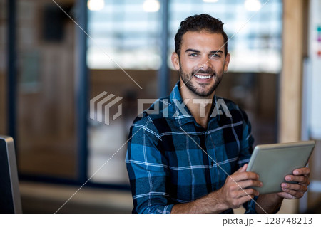 Bearded man wearing plaid shirt sitting at desk in office meeting room holding tablet while smiling 128748213