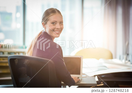 Businesswoman smiling and working at office desk by large windows with laptop and blueprints 128748218