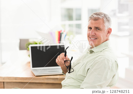 Senior man sitting at home office holding eyeglasses and smiling by laptop and folders, copy space 128748251