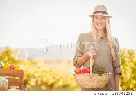 Woman holding basket with tomatoes and herbs in sunlit farm field wearing straw hat, copy space 128748252