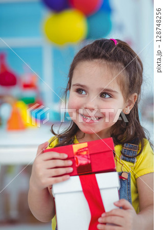 Preschool girl holding two-tiered gift box at party room, glancing right among balloons and toys 128748256
