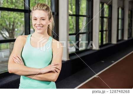 Woman standing inside sports facility, crossing arms, smiling near running track and glass windows Woman standing inside sports facility, crossing arms, smiling near running track and glass windows 128748268