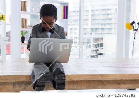 African American boy in formal suit sitting at desk using silver laptop in office with plant 128748292