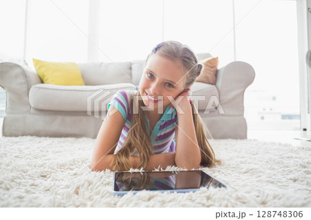 School-age girl lying on plush white rug in bright living room gazing at tablet 128748306