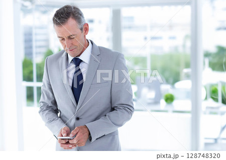 Male professional holding smartphone while standing in modern office by desk, monitor, potted plant 128748320