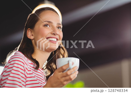Woman in twenties wearing red-striped tee smiling and holding white ceramic mug at cafe, copy space 128748334