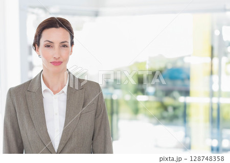 Mid adult woman wearing gray blazer standing in office lobby near glass panels revealing greenery Mid adult woman wearing gray blazer standing in office lobby near glass panels revealing greenery 128748358