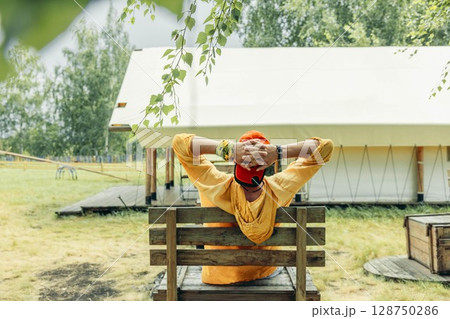 man in orange cap, yellow hoodie leaning back on wooden bench with hands behind head in front of glamping tent in forest meadow on summer day, travel eco tourism slow lifestyle retreat 128750286