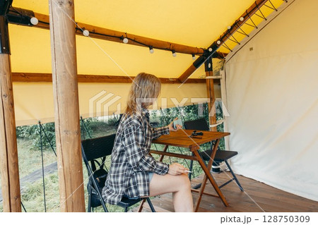 woman sitting inside glamping tent holding cigarette and phone on wooden chair at rustic table with casual outfit under yellow canopy, outdoor travel, countryside lifestyle, digital detox 128750309